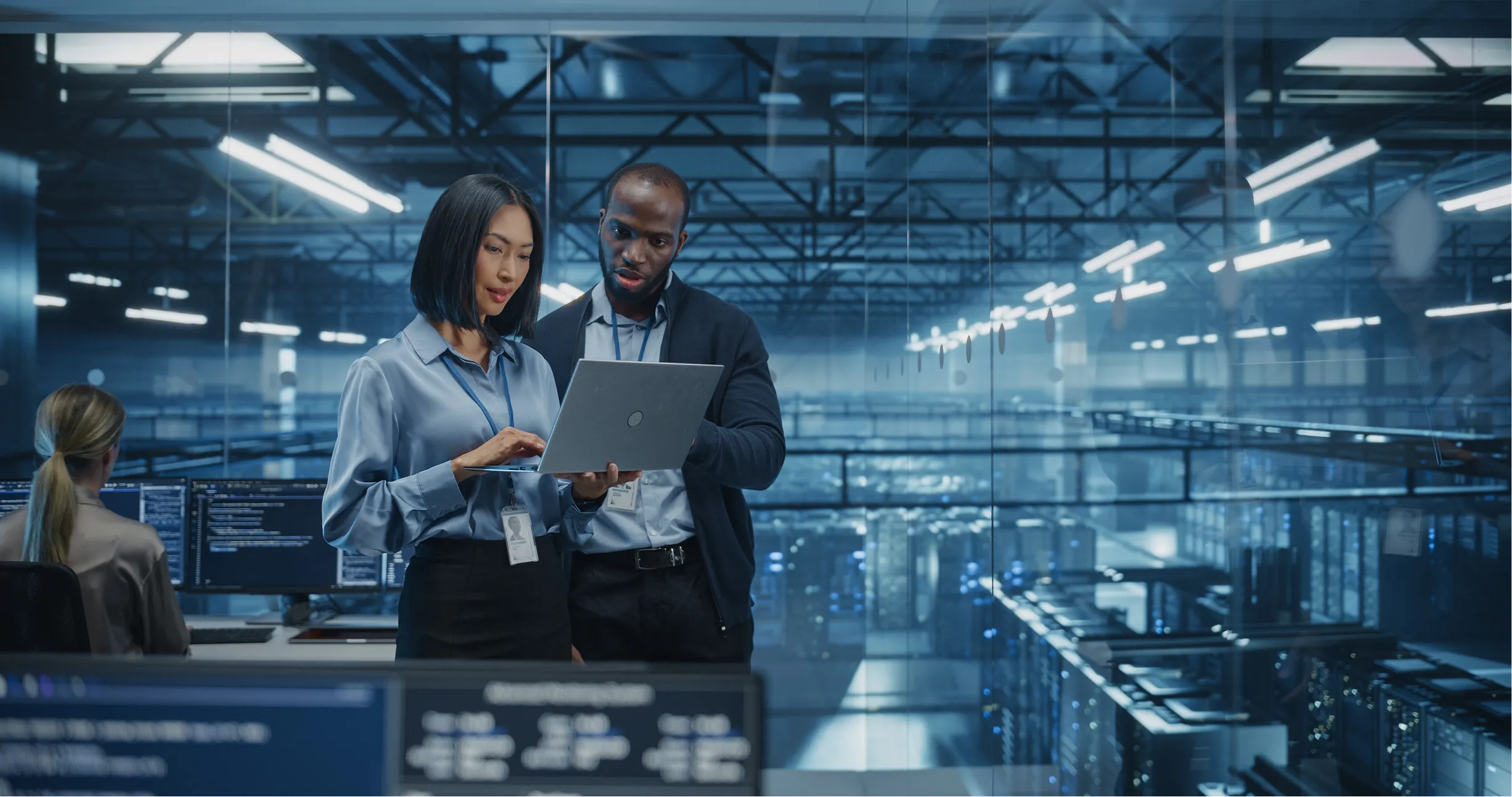 Two IT professionals stand in a modern server room, reviewing information on a laptop. They are surrounded by glass walls and rows of server racks. One person gestures toward the screen; another person works at a desk in the background.
