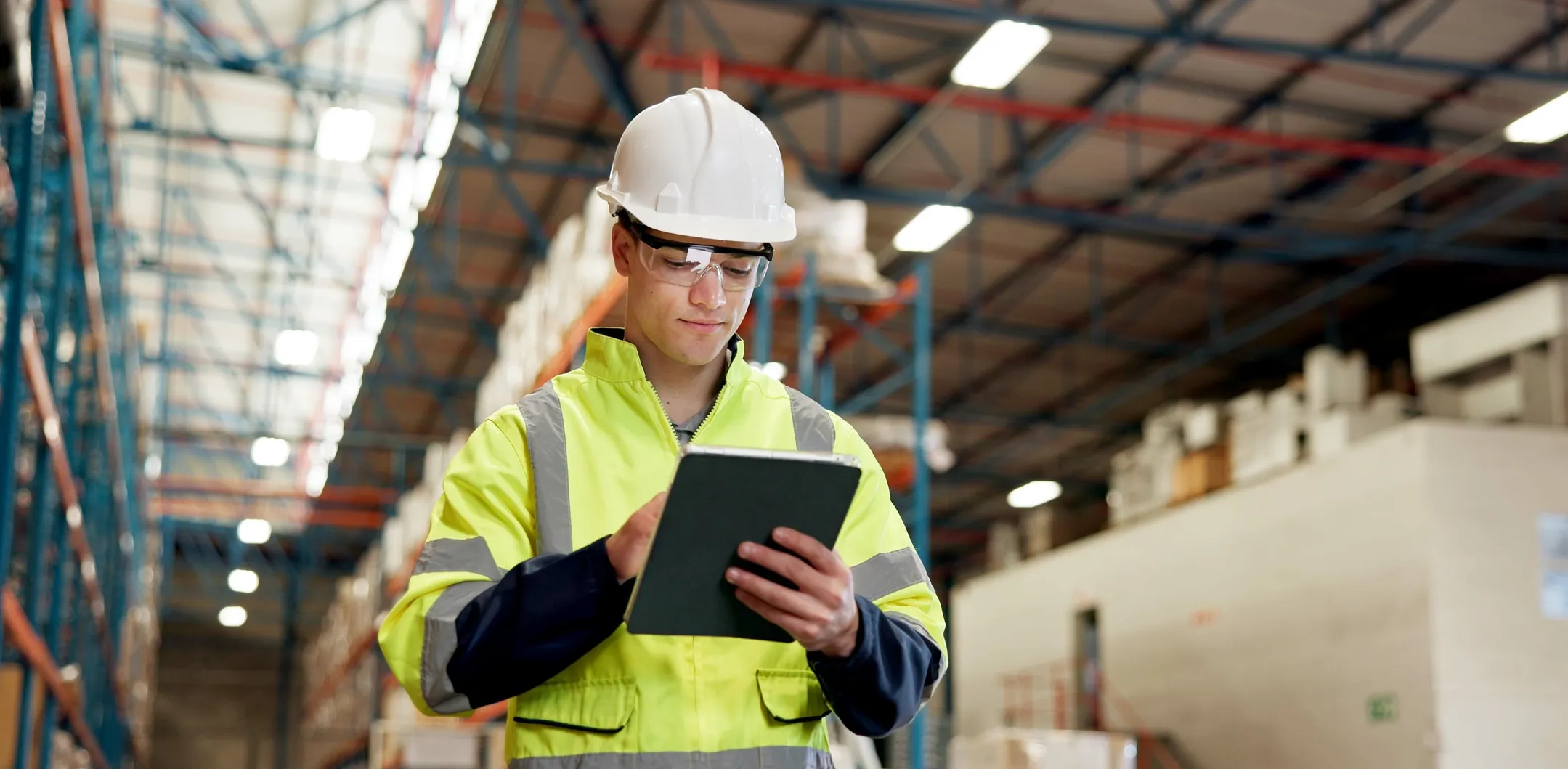 A worker wearing a white hard hat, safety glasses, and a fluorescent yellow jacket uses a tablet in a large warehouse with high shelves and boxes. The ceiling has exposed beams and bright lights, and the background shows industrial storage areas.