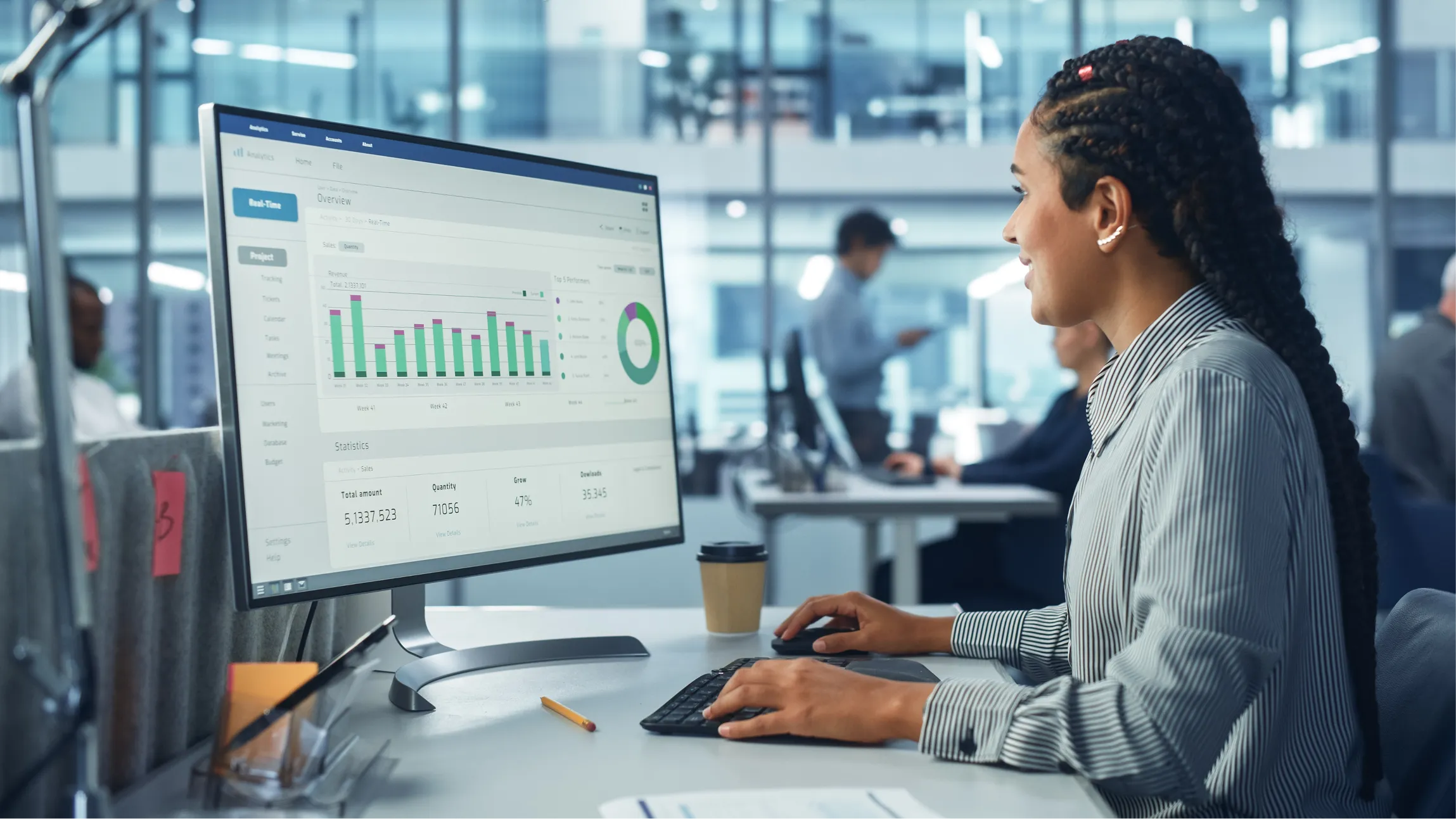 A woman sits at a desk in an office, looking at a large computer monitor displaying graphs and charts. She types on a keyboard and smiles. A coffee cup sits nearby. The office background is modern and bright, with blurred coworkers and glass walls.