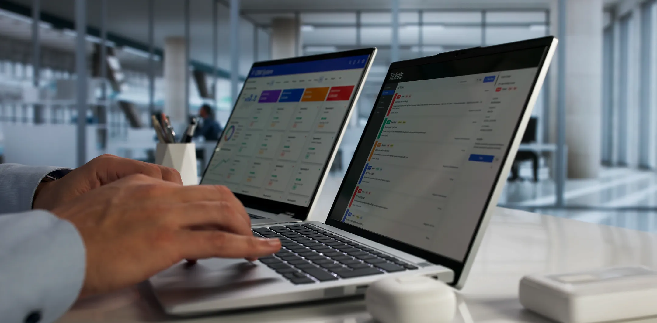 A person types on a laptop at a modern office desk, with a second open laptop beside them. Both screens display dashboards with colorful charts, graphs, and tables. Office supplies and a bright, glass-walled workspace are visible in the background.