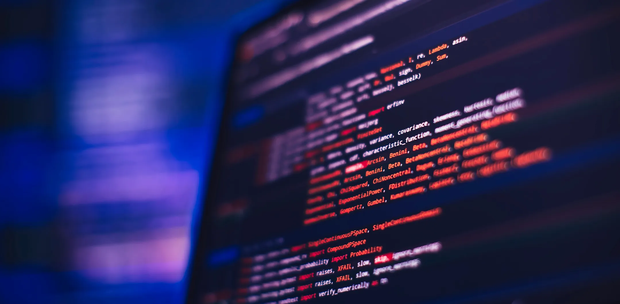A close-up of a computer screen displaying lines of colorful code in a text editor, with syntax highlighting in red, orange, purple, and white on a dark background. The image has a blurred, blue-toned background, suggesting a tech-focused environment.