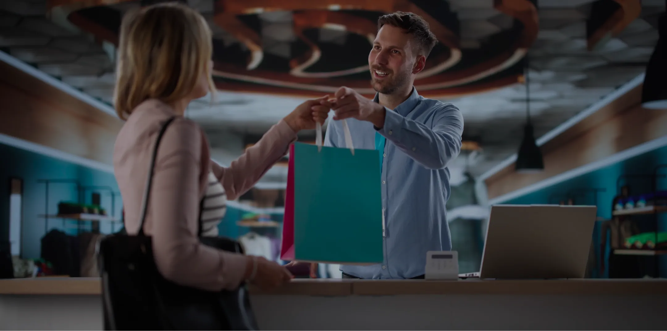 A smiling man behind a counter hands a turquoise shopping bag to a woman in a light jacket. The woman faces away, holding a black purse. A laptop and small card reader sit on the counter. The background shows modern decor and shelves.