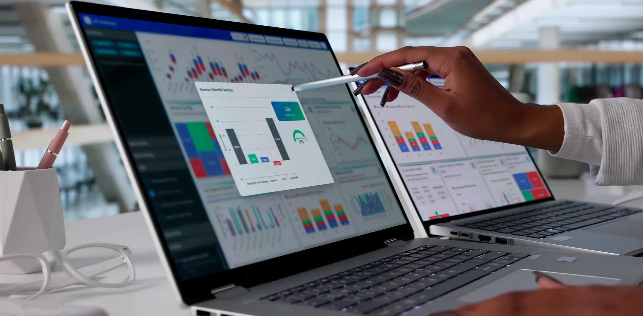 A person points a pen at a laptop screen displaying various colorful data charts and graphs. Another laptop with similar charts is visible in the background. The setting appears to be a modern office with bright natural light.