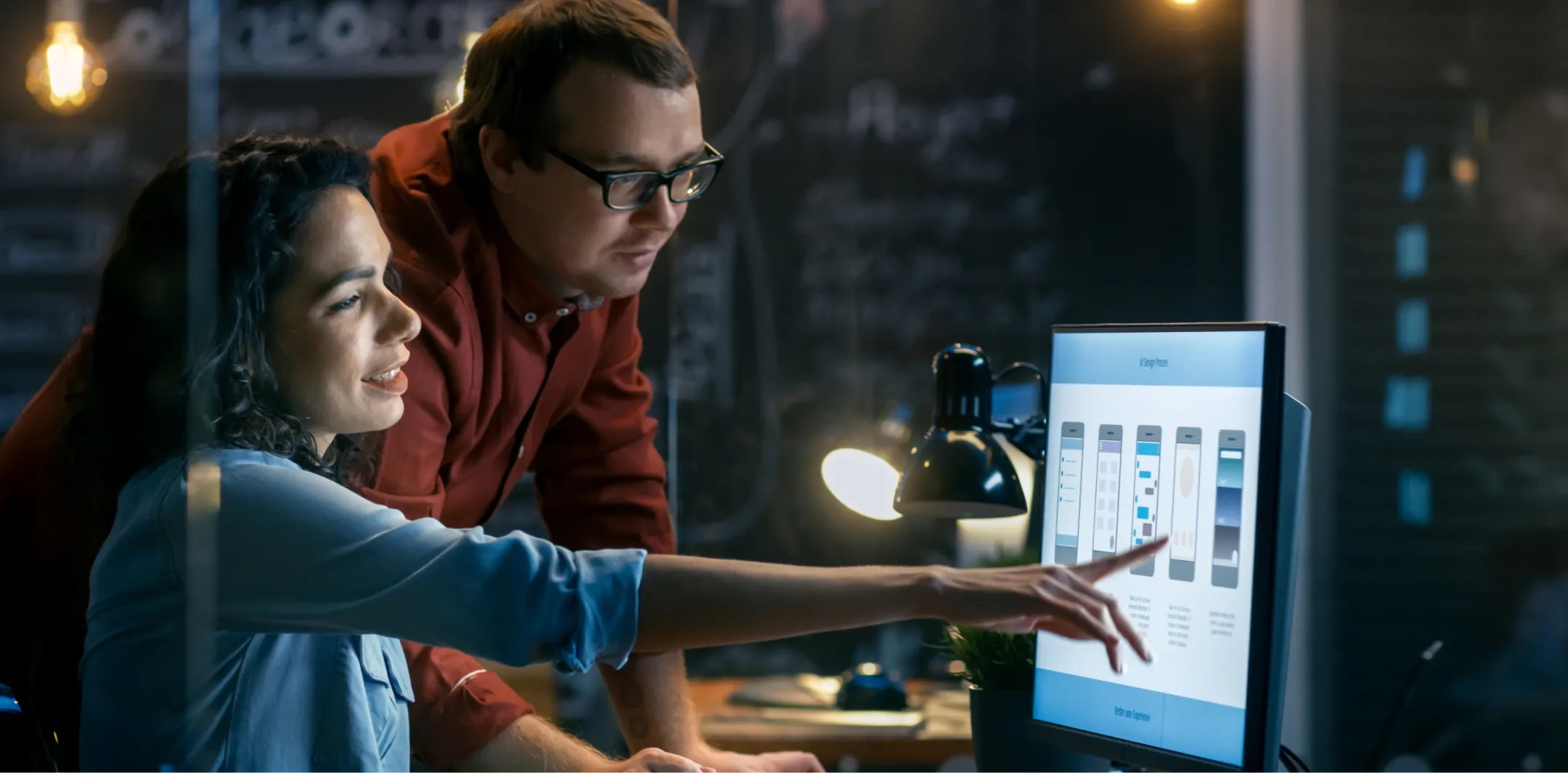 A woman sits at a desk, smiling and pointing at a desktop monitor displaying interface designs, while a man in glasses stands beside her, looking at the screen. The room is dimly lit with a desk lamp and a chalkboard wall in the background.