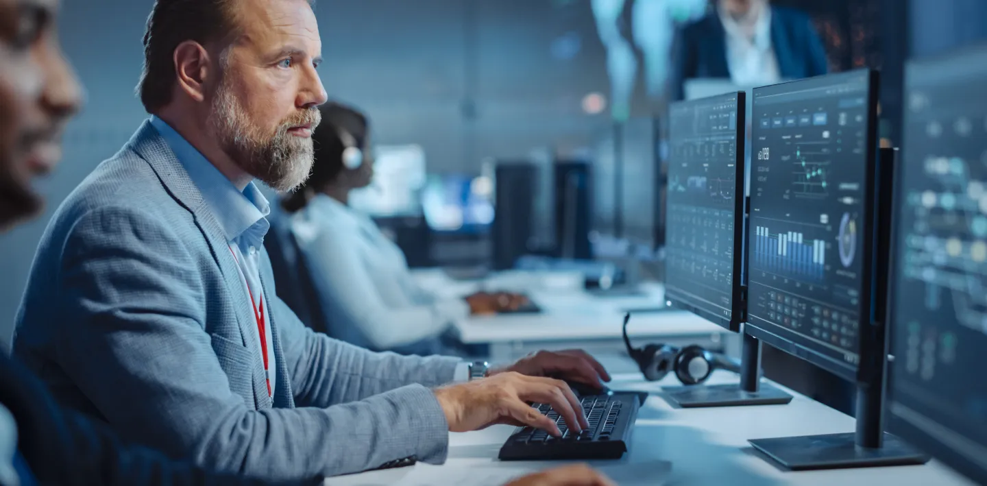 A man in a blue suit sits at a desk, typing on a keyboard and looking at multiple computer monitors displaying data and graphs. He is in a busy, modern office with other professionals working at computers in the background.