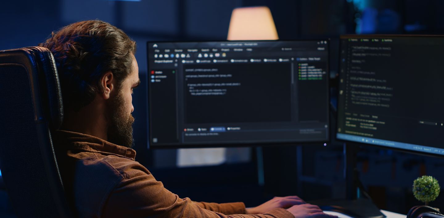 Man in a dimly lit room working on a computer with code on dual monitors, a lamp in the background.