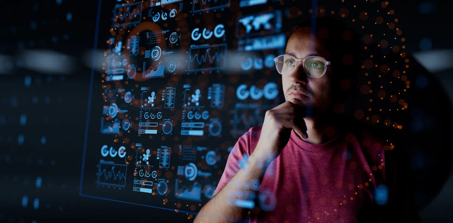 A man in glasses and a red shirt looks thoughtfully at a transparent screen filled with colorful, futuristic data visualizations, charts, and graphs in a dark setting, suggesting data analysis or technological problem-solving.