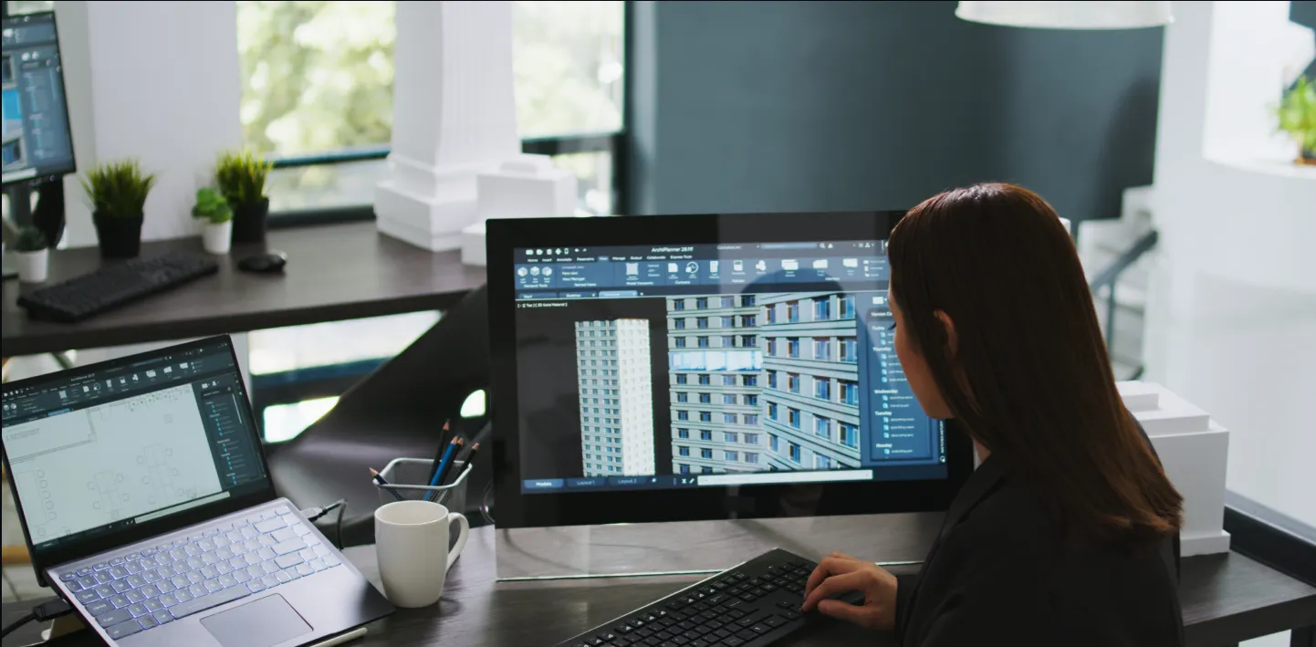 A person with long brown hair sits at a desk working on a desktop computer, displaying architectural 3D building models. A laptop, coffee mug, pen holder, and potted plants are on the desk. Large windows and architectural models are in the background.