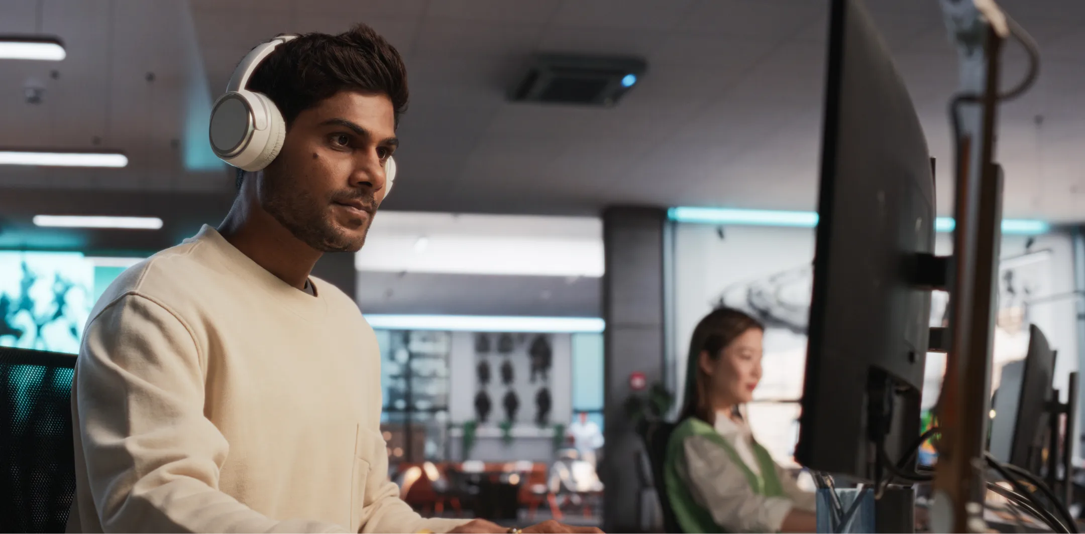 A man wearing white headphones and a light sweater sits at a desk using a computer in a modern office. In the background, a woman is also working at a desk. The office is spacious, with artwork and bright lighting.