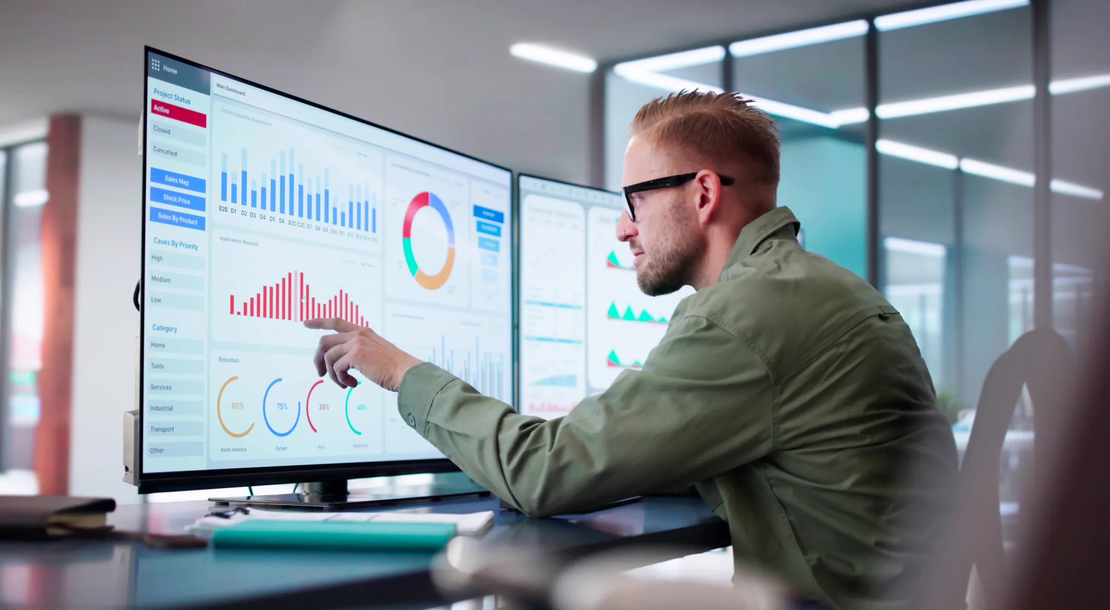 A man with glasses and a beard sits at a desk in a modern office, analyzing colorful charts and graphs displayed on a large computer monitor. He points at a red bar graph, focusing on data related to business analytics and performance metrics.