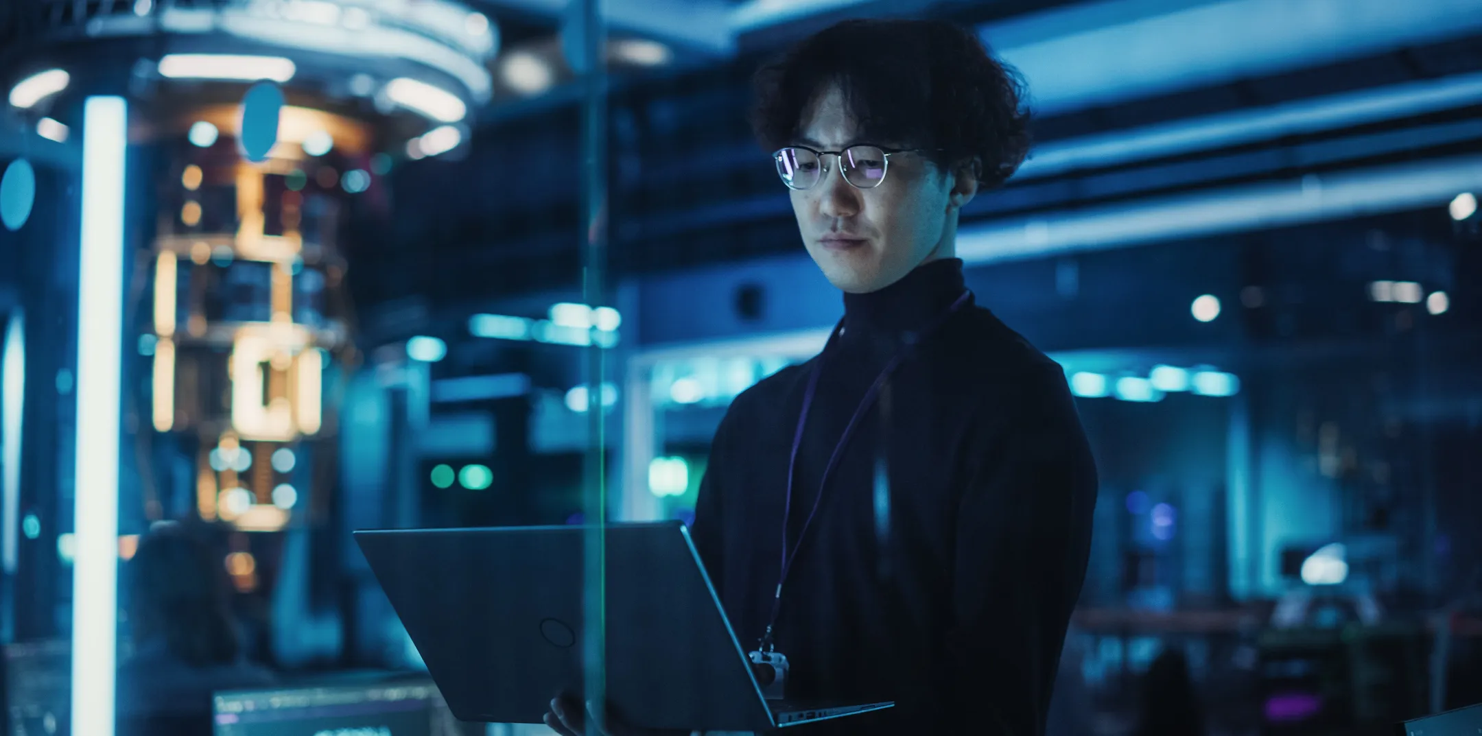 A person with glasses and curly dark hair stands in a futuristic, blue-lit server room or data center, looking at an open laptop. They wear a black turtleneck and lanyard, surrounded by glowing technological equipment and reflections.
