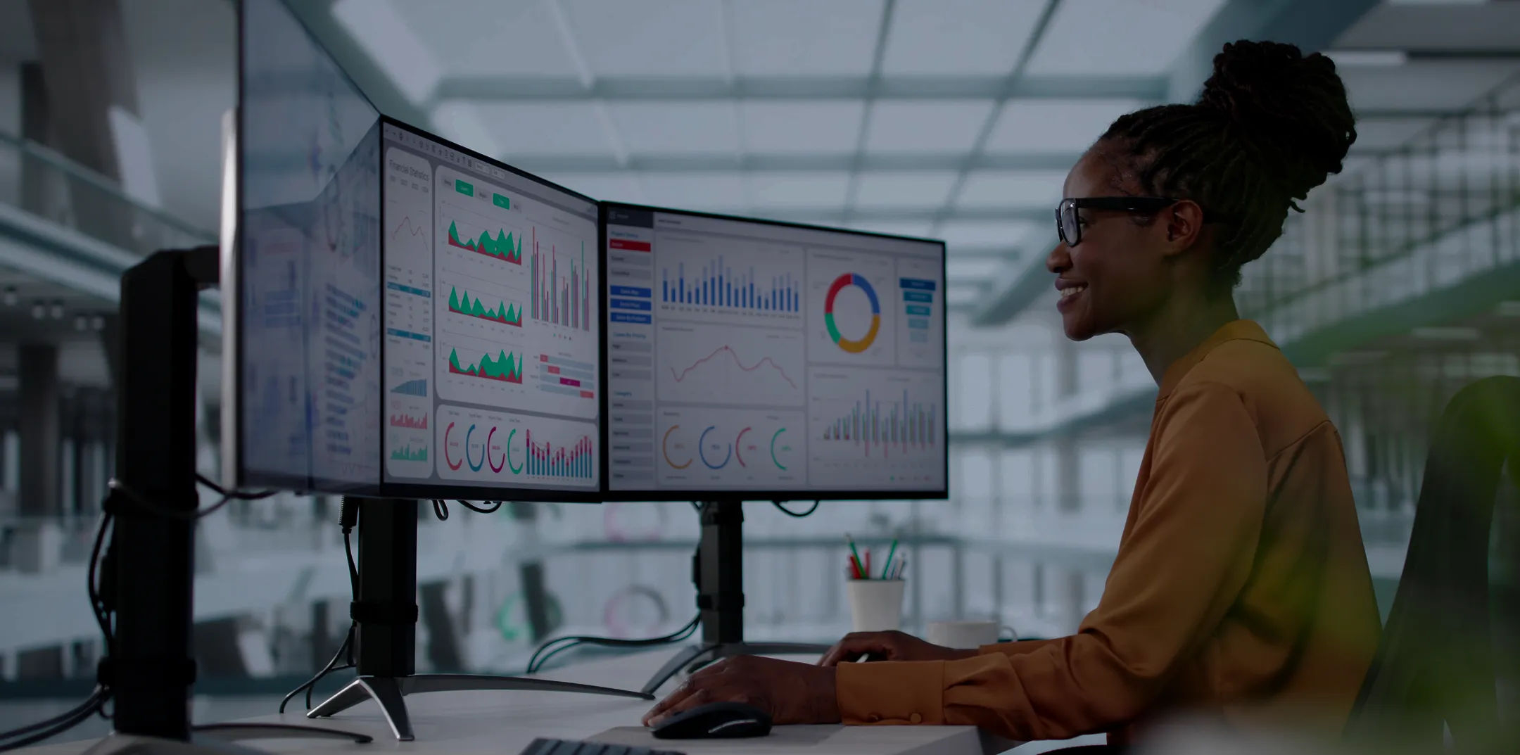 A woman with glasses sits at a desk in a modern office, working on two large computer monitors displaying colorful data charts and graphs. She is focused and smiling, using a mouse and keyboard, with a bright, open workspace in the background.