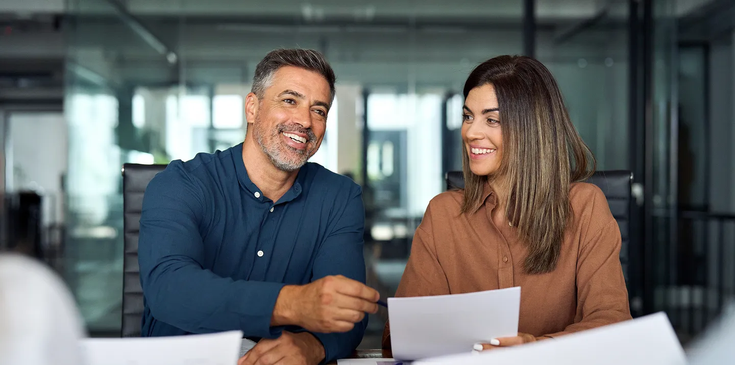 A man and a woman sit at a conference table in a modern office, smiling and holding papers. The man, with salt-and-pepper hair and beard, wears a blue shirt; the woman, with long brown hair, wears a brown blouse. Glass walls and office furniture are visible behind them.