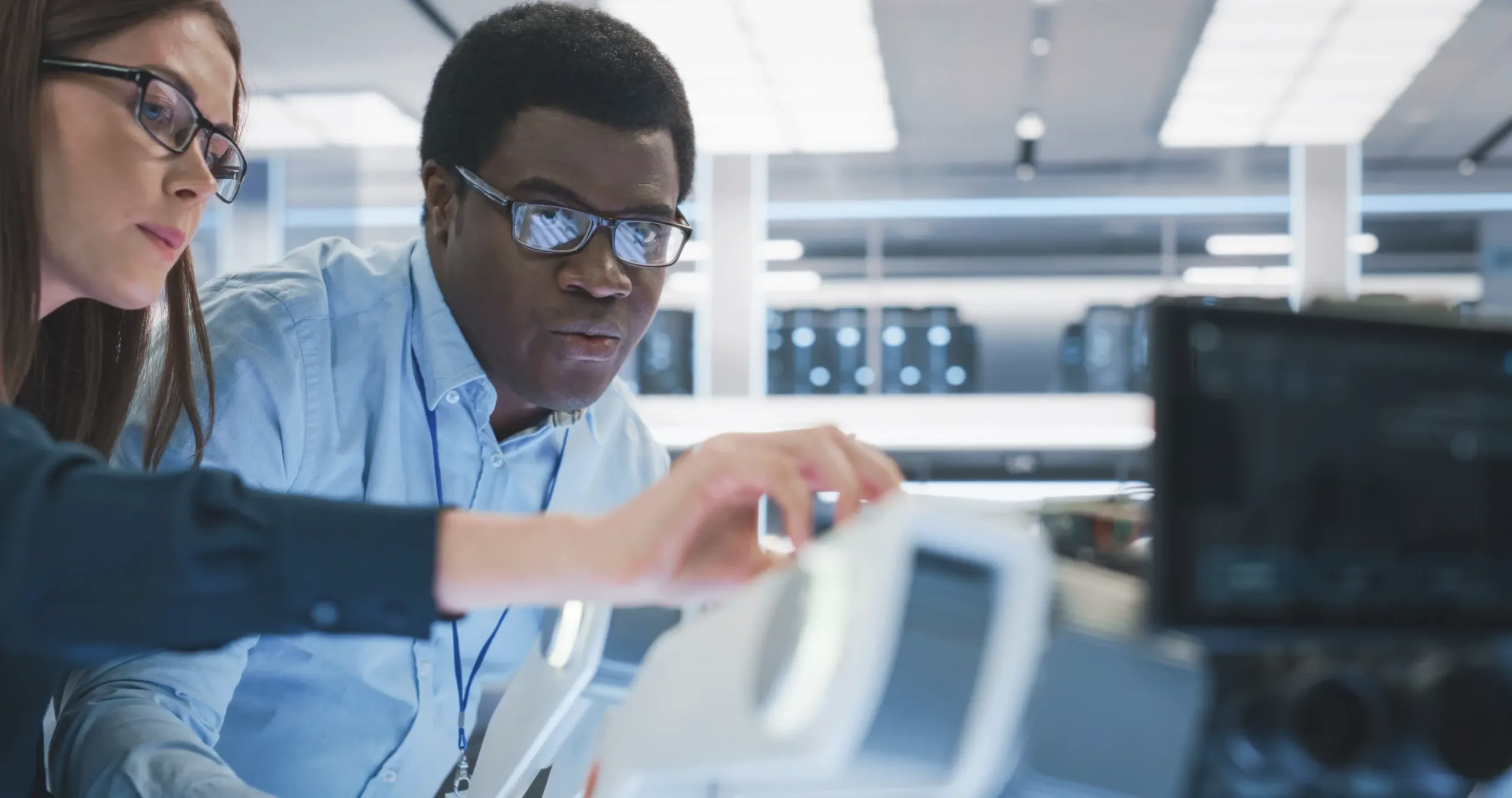 Two people wearing glasses examine technical equipment in a bright, modern lab. The man in focus looks closely at a device while the woman points toward it. Computer servers and lab lights are visible in the background, suggesting a high-tech environment.