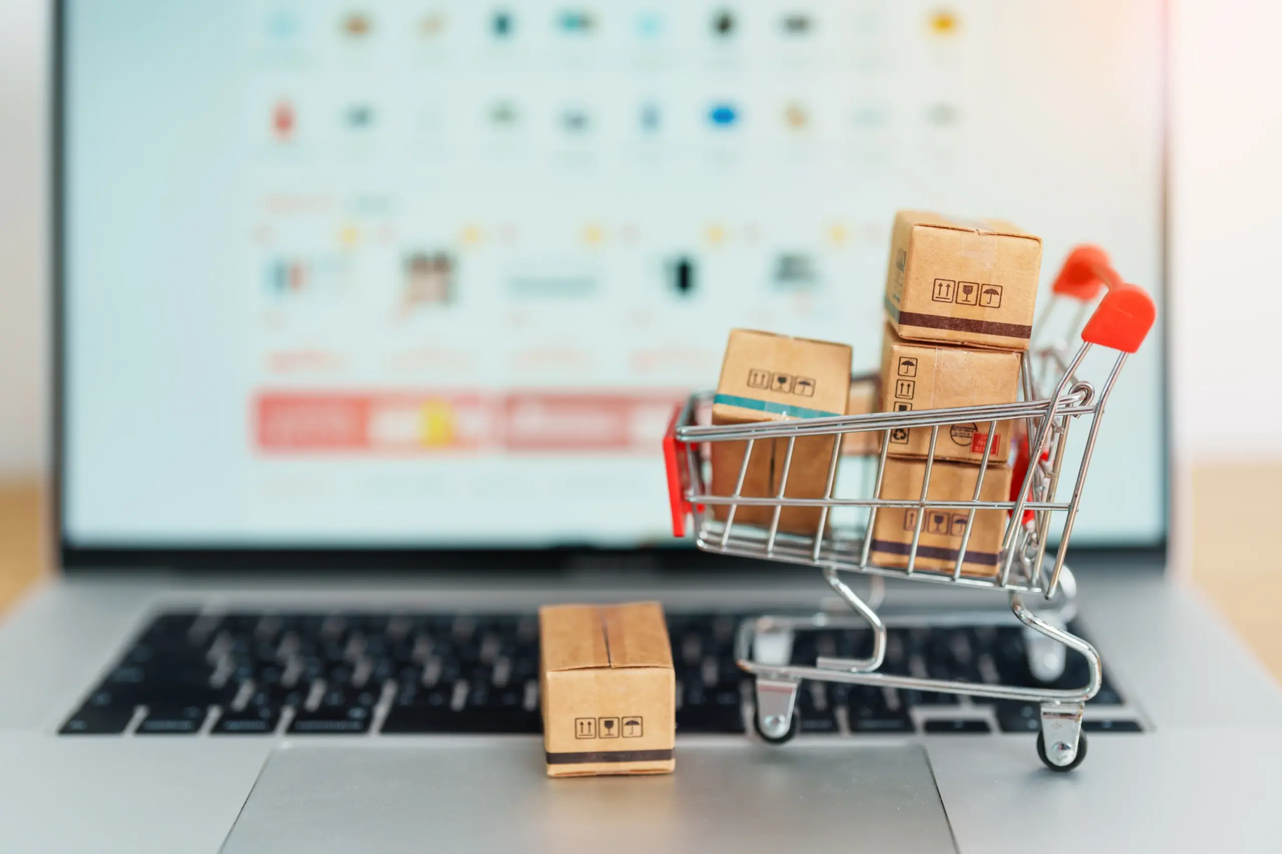 A miniature shopping cart filled with small cardboard boxes sits on a laptop keyboard. The laptop screen in the background displays a blurred e-commerce website, symbolizing online shopping and parcel delivery.