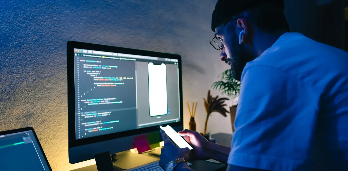 A man wearing glasses and a cap sits at a desk in a dim room, viewing code on a large computer monitor. He holds a smartphone and wears wireless earbuds. Desk items include a keyboard, mouse, sticky notes, and a plant in the background.