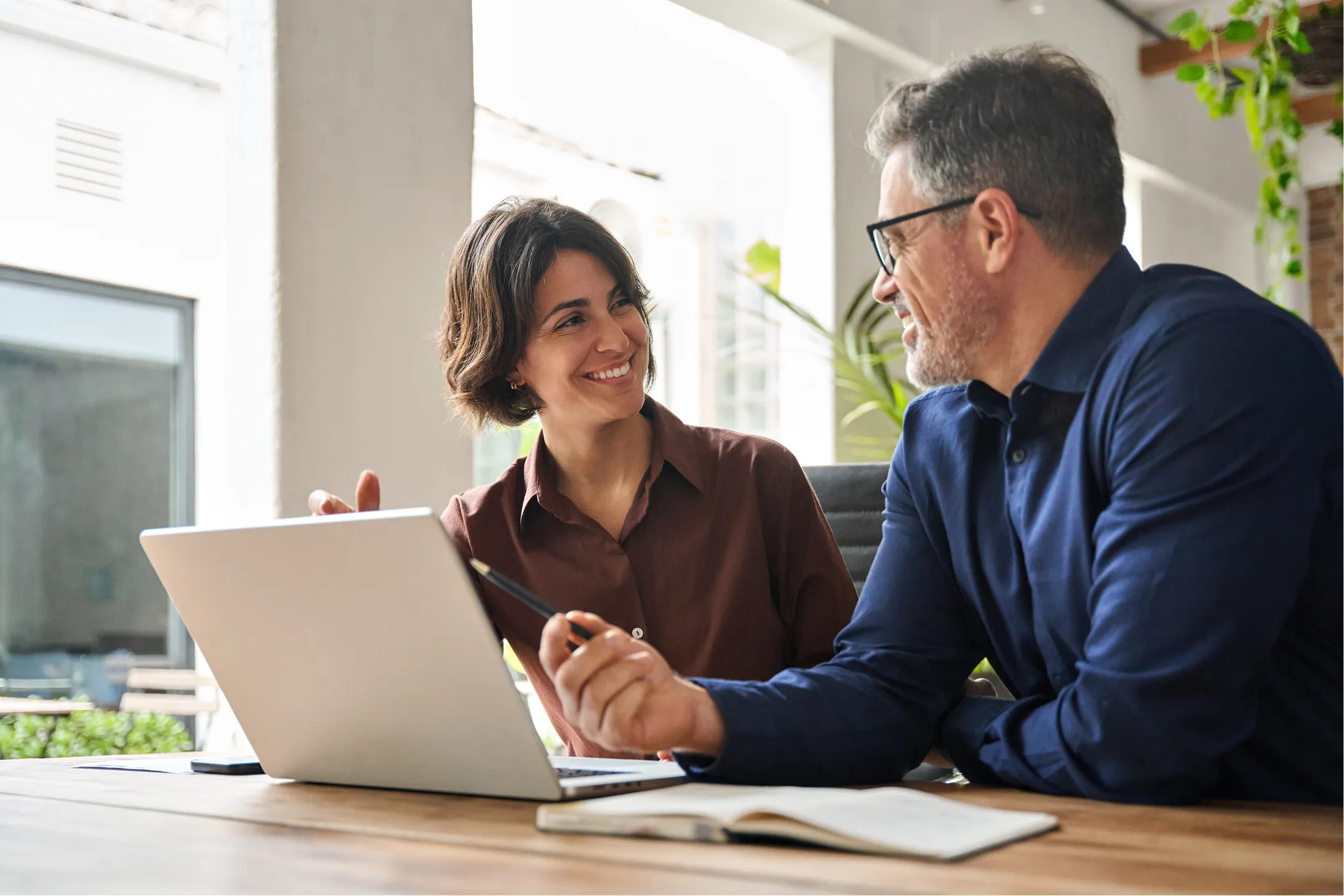 A woman and a man sit at a table in a bright office, smiling and talking. The woman has short brown hair and wears a brown blouse, while the man has gray hair, glasses, and a navy shirt. A laptop and open notebook are in front of them.