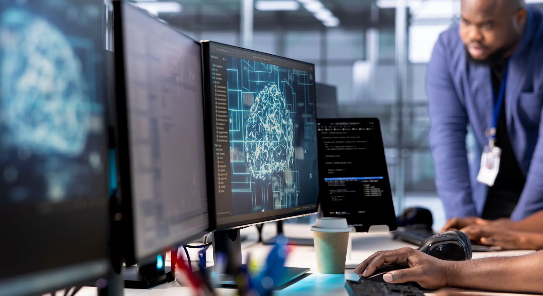 A person works at a desk with multiple computer monitors displaying a digital brain and code. Another person stands nearby, watching. The office setting is modern, with bright lighting and glass walls. A coffee cup and headphones are on the desk.