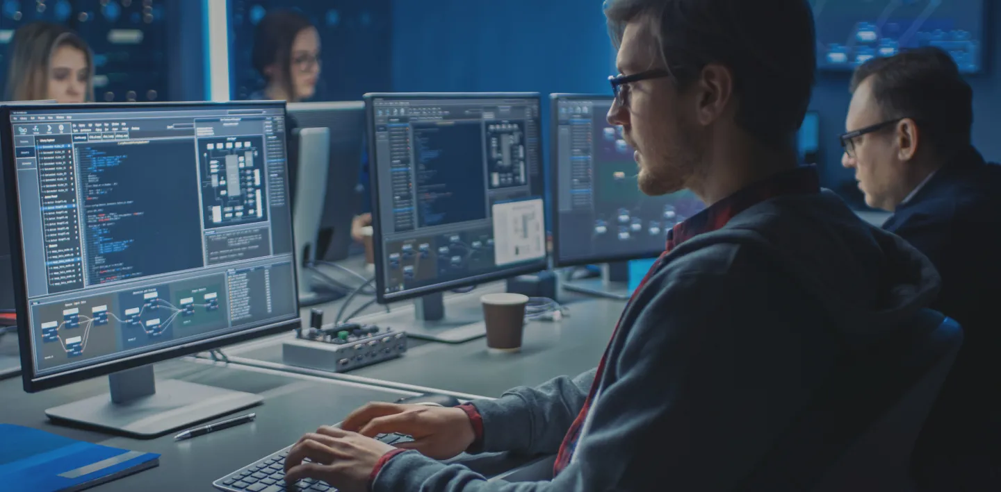 A man in glasses works at a computer with multiple monitors displaying code and data. Other people are seated at desks in the background, also working on computers in a modern, dimly lit office with blue lighting.