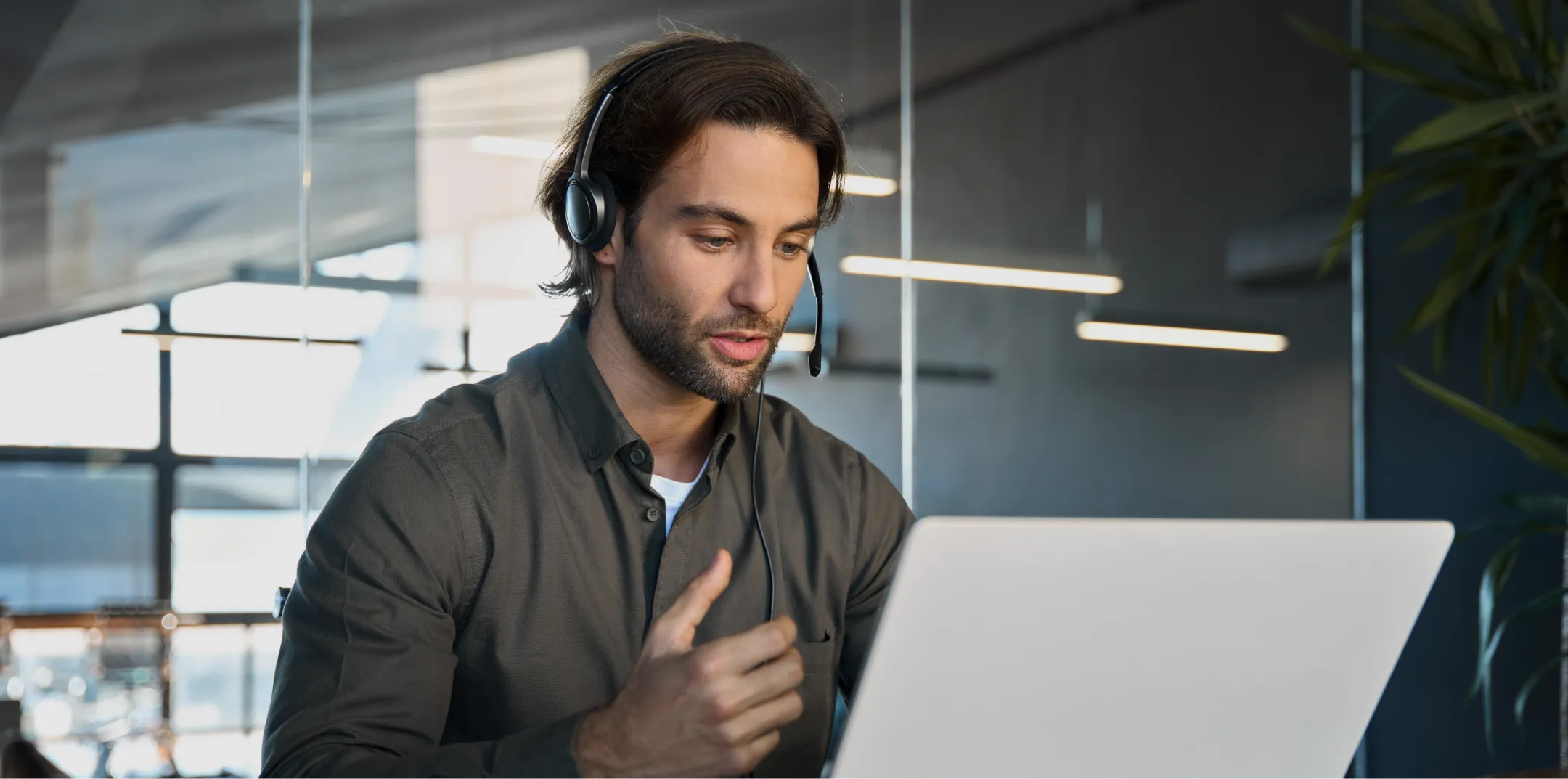 A man with long brown hair and a beard, wearing a headset and gray shirt, sits at a desk using a laptop in a modern office with glass walls. He appears focused, gesturing with his hand as if speaking during a video call or online meeting.