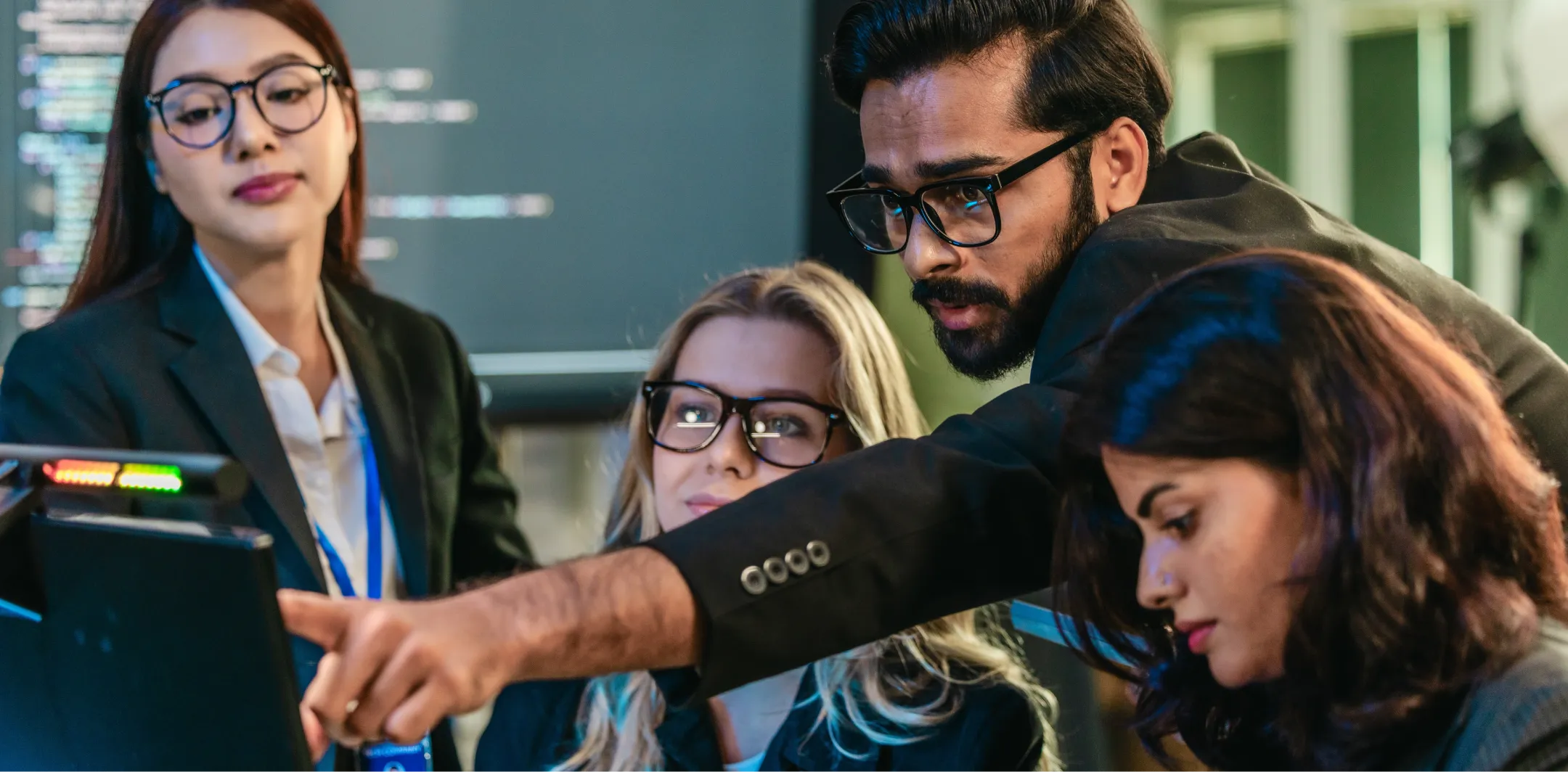 Four professionals gather around a computer monitor in an office setting. One man points at the screen while the three women, all wearing glasses, focus intently. There is code visible on a screen in the background, suggesting a tech or programming context.
