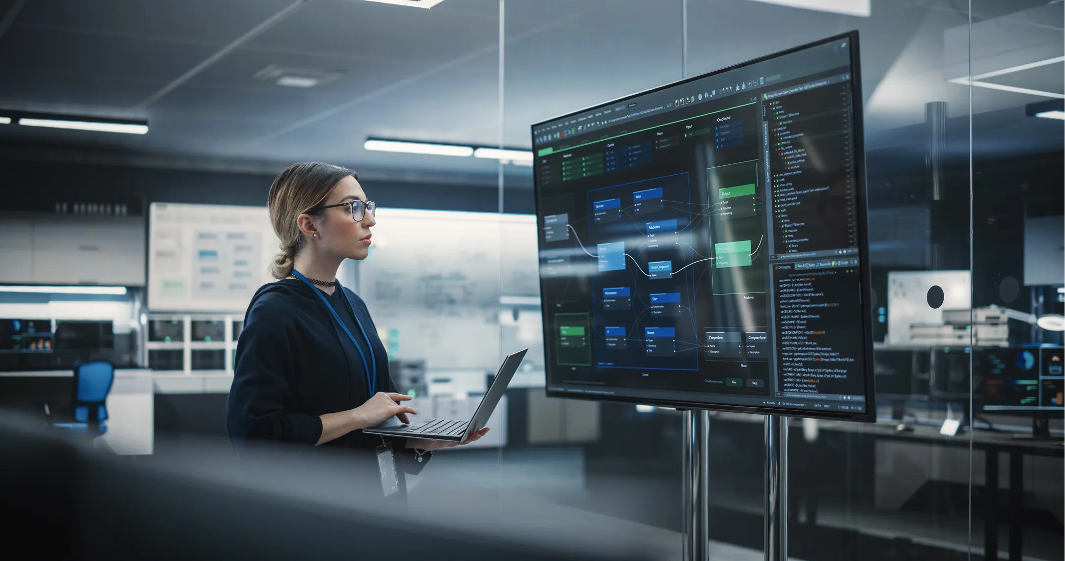 A woman with glasses and a ponytail holds a laptop, analyzing flowcharts and code on a large screen in a modern, high-tech office with glass walls and sophisticated electronic equipment in the background.