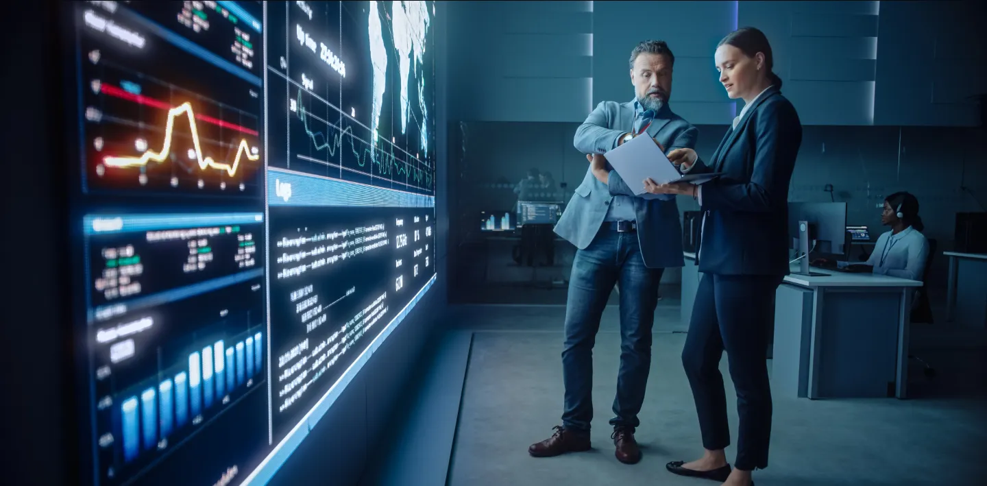 Two professionals in business attire discuss documents in a modern office. Large digital screens display charts, graphs, and data visualizations. Another person works at a computer in the background. The setting is high-tech and data-driven.