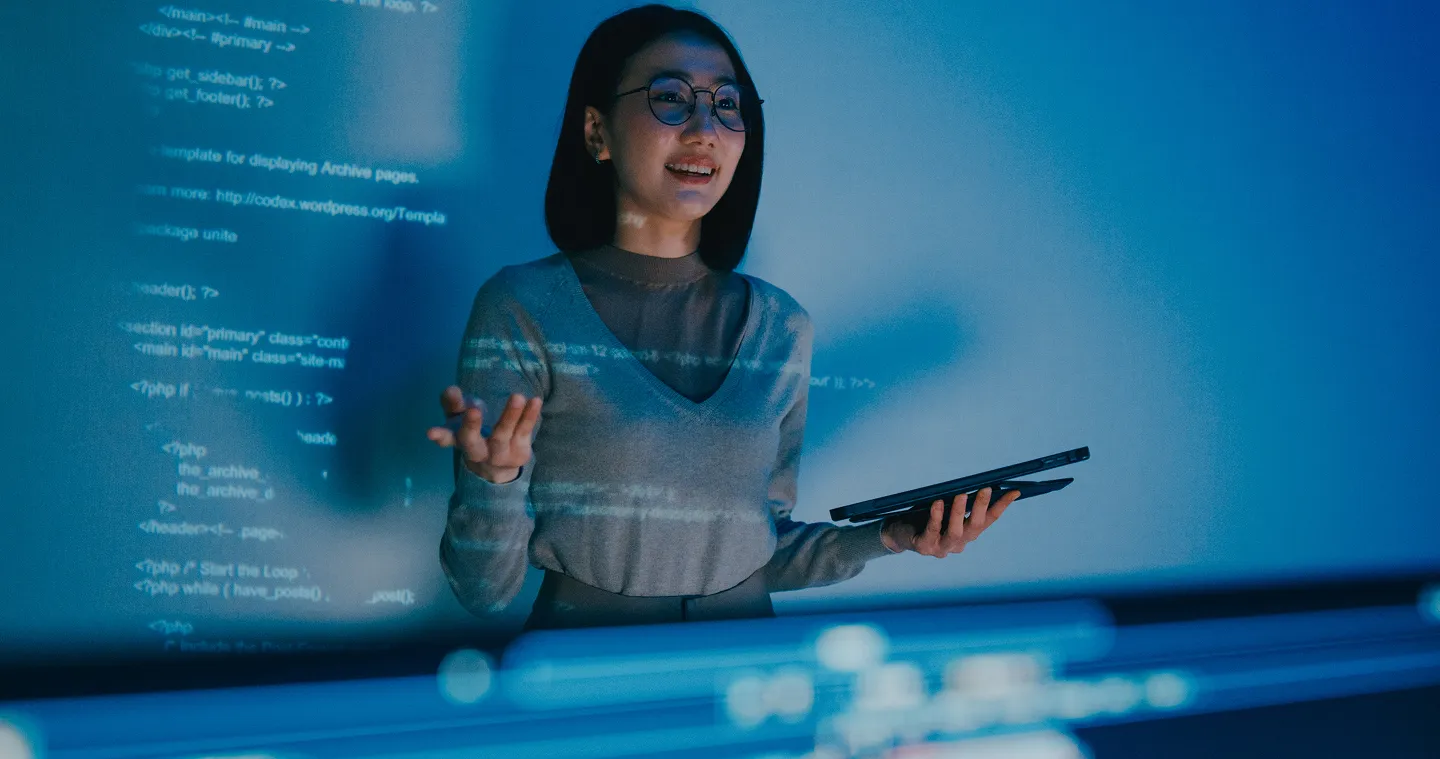 A woman with glasses stands in front of a screen displaying lines of code, holding a tablet and gesturing as if presenting or explaining something. The blue lighting and digital projections create a high-tech, modern atmosphere.