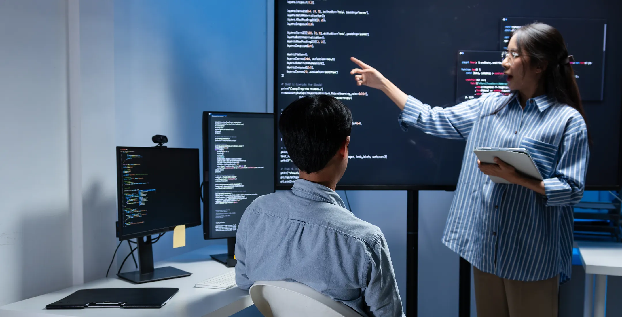 A woman holding a tablet points at code on a large screen, explaining to a seated man in front of multiple monitors displaying code in a modern office. The room is dimly lit, creating a focused and professional atmosphere.